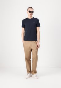 Kék crew-neck póló, bézs chinos nadrág és fehér sportcipő. A modellen fekete napszemüveg van, rövid haja van, és egy sima háttér előtt áll.