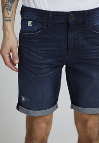 Pantalones cortos de denim oscuro con una textura desvanecida, dobladillos enrollados, banda interior en contraste y un pequeño parche de logo en el bolsillo frontal.