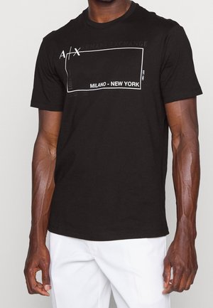 Mężczyzna w czarnej koszulce Armani Exchange z białym prostokątem i napisem "MILANO - NEW YORK" oraz białych spodniach na neutralnym tle.