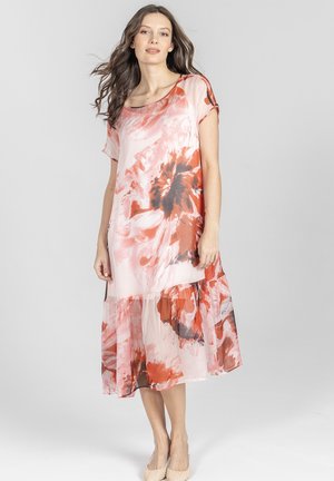 Kobieta ubrana w zwiewną sukienkę do kolan z pomarańczowo-czarnymi i białymi abstrakcyjnymi wzorami kwiatowymi, stojąca na jednolitym jasnym tle.