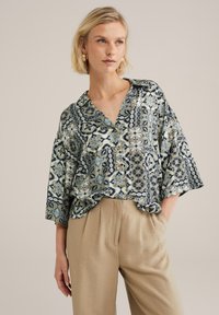 Ovratna, ohlapna srajca v modro-zeleni cvetlični vzorcu z gumbi in kratkimi sleeves, v kombinaciji z bež teksturiranimi hlačami.