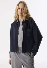 Marineblauwe overhemd met knopen en een borstzak, gemaakt van stoffig materiaal. Daaronder een lichtgrijs T-shirt met korte mouwen en bedrukte tekst. Grijze broek.