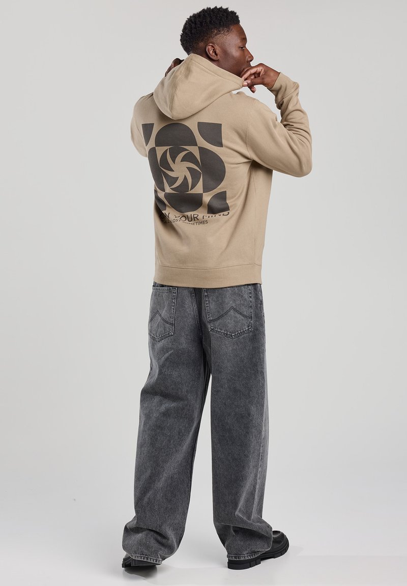 Beige hoodie with a black swirl design on the back, paired with gray baggy jeans. Material appears soft, featuring a relaxed fit and drawstring hood.