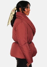 Rød boblejakke med bred krage og belte i midjen. Har glatt, polstret tekstur og sideprofil av et lyst grønt silkeskjerf i håret.