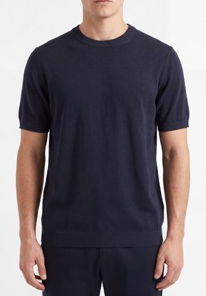 Uomo che indossa un maglione a maniche corte blu navy semplice e pantaloni abbinati, in piedi con le braccia rilassate ai lati su sfondo bianco.