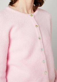 Šviesiai rožinė megztinė, pagamintas iš minkštos medžiagos, su apvaliu kaklu ir šešiais daugiabutų mygtukais priekyje.