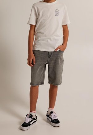 Witte katoenen T-shirt met korte mouwen en grafische print. Grijze denim shorts, knielang. Zwart-witte sneakers met rode accenten.