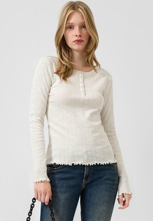 Junge Frau mit blondem Haar, die ein cremefarbenes Langarmoberteil mit Knöpfen und blaue Jeans trägt, hält eine schwarze Kettenhandtasche vor einem schlichten Hintergrund.