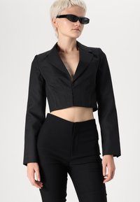 Svart kort blazer med lange ermer, strukturert passform og vertikale sømdetaljer, sammen med høy i livet svarte bukser. Strukturert stoff.