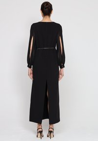 Czarna długa sukienka z przezroczystymi długimi rękawami, rozcięciem z tyłu i dopasowanym pasem. Gładka tekstura i elegancka sylwetka.