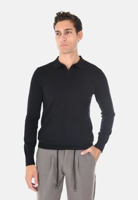 Polo a maniche lunghe nera con scollo a V e polsini a coste, realizzata in un tessuto liscio, abbinata a pantaloni chiari con cordoncino.