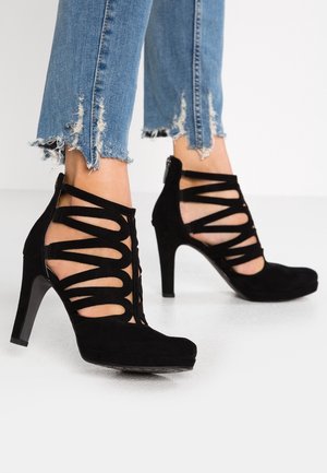 Femme portant un jean bleu à ourlet déchiré et des chaussures noires à talons hauts avec plusieurs lanières découpées et une fermeture éclair au dos.