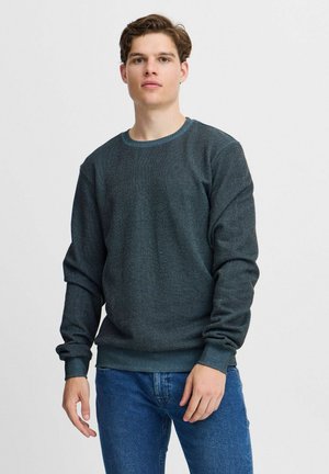 Sweatshirt vert foncé texturé avec un col rond et des poignets côtelés. Associé à un jean bleu, présenté sur un fond neutre.