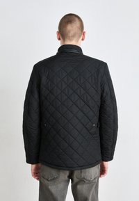 Μαύρο επενδυμένο μπουφάν με διαμαντέ μοτίβο, ψηλό γιακά και κουμπιά με κλίπ. Διαθέτει μακριά μανίκια και ίσια γραμμή. Φοριέται πάνω από γκρι παντελόνι.