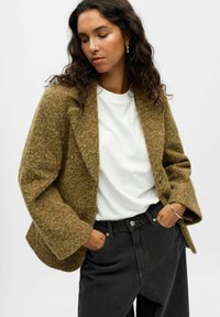 Olijfgroene, gestructureerde wolmix blazer met een enkele knoopsluiting en brede mouwen, gedragen over een licht t-shirt en donkere jeans.