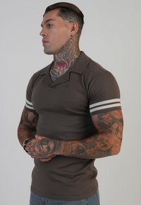 Hnědý polo triko s límečkem, které zdobí šedé pruhy na krátkých rukávech. Materiál vypadá texturovaně, s přiléhavým střihem, který zvýrazňuje trup.