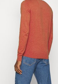 Pull en tricot couleur rouille avec manches longues, col rond et poignets côtelés. Associé à un jean bleu avec un design simple sur la poche arrière.