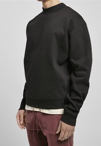Sudadera negra de cuello redondo con ribete en el cuello y los puños. Textura suave, corte relajado. Usada sobre una camisa de color claro y combinada con pantalones burdeos.