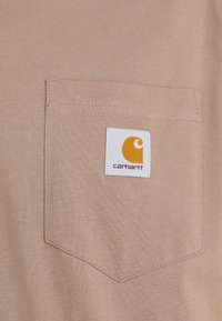 Beige t-paita, jossa on tilkkutasku, jossa on valkoinen etiketti, jossa on Carhartt-logoa oranssina ja ruskeana. Pehmeä kangas, jossa on sileä rakenne.