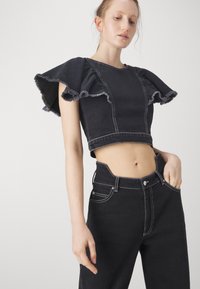 Svart denim crop-top med volangermar, rå kanter och kontrasterande sömmar. Kombinerad med högmidjade svarta denimbyxor, även de med sömnadsdetaljer.