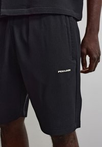 Gros plan d'une personne portant un short noir côtelé avec une poche latérale et le logo blanc "PEGADOR" sur la cuisse gauche.