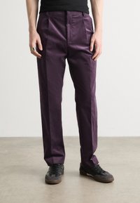 Pantalons violets en tissu lisse, avec une taille haute et des plis à l'avant. Portés avec des baskets noires, ils ont une coupe ajustée.