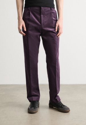 Pantalons violets en tissu lisse, avec une taille haute et des plis à l'avant. Portés avec des baskets noires, ils ont une coupe ajustée.