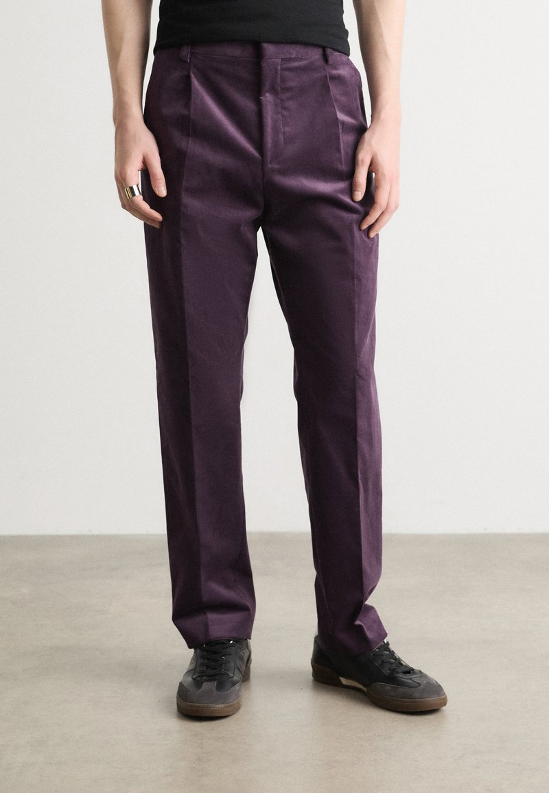 Pantalons violets en tissu lisse, avec une taille haute et des plis à l'avant. Portés avec des baskets noires, ils ont une coupe ajustée.