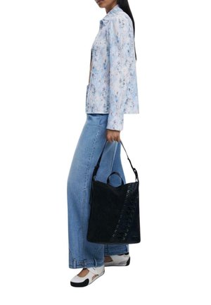 Zwarte suède handtas met decoratieve vetersluiting, gecombineerd met een lichtblauw shirt met bloemmotief en wijde denim jeans.
