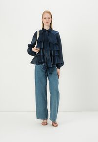 Blouse en soie bleu marine avec des couches volantées, col boutonné et détail en nœud, associée à un jean en denim bleu clair ample et des sandales beiges.