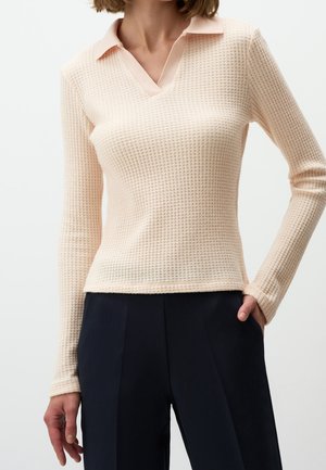 Femme portant un haut beige texturé à manches longues avec col, pantalon noir taille haute, main dans la poche.