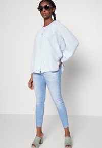 Top de tecido azul claro e enrugado com decote redondo e botões frontais, combinado com jeans skinny de lavagem clara e mules de salto verdes. Óculos de sol pretos.