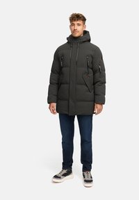 Polstret sort jakke med hætte, med flere lynlåslommer og en tekstureret overflade. Båret med blå jeans og sneakers med sorte såler.
