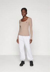 Bež dugi rukav s okruglim izrezom i logotipom, upareno s bijelim trenirkama i crnim tenisicama. Glatka tekstura, ležerni kroj.