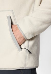 Fleece-genser i off-white med grå kant. Har en frontlomme og justerbar snor. Myk tekstur med synlige sømdetaljer.