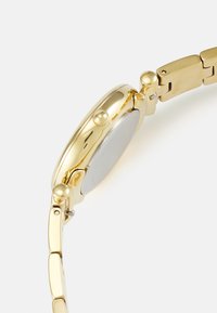 Goldfarbene Armbanduhr mit runder Gehäuseform, glatter Oberfläche und einem miteinander verbundenen Armband. Enthält seitliche Drücker und ein poliertes Zifferblatt aus Metall.