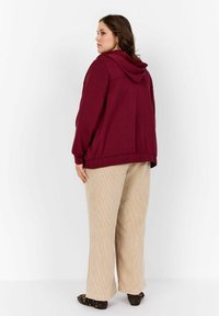 Sudadera burdeos con capucha ajustable, combinada con pantalones de tiro ancho de color beige y zapatos oscuros. El tejido parece suave y casual.