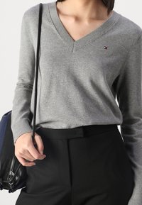 Maglione grigio a scollo a V con maniche lunghe, polsini a costine e un piccolo dettaglio logo. Abbinato a pantaloni neri su misura e una borsa a tracolla.