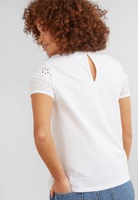 Haut en coton blanc avec manches courtes brodées de dentelle, détail dos en forme de trou de serrure et coupe décontractée. Porté avec un bas en denim bleu.