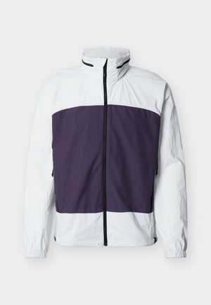 Balta jaka ar violetu priekšpuses paneļu krūtīs, augstu apkakli, pilnu melnu priekšējo rāvējslēdzi un elastīgiem aproču galiem uz piedurknēm.