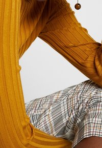 Senapsgul ribbad tröjärm med texturerade detaljer, i kombination med en rutigt tyg i vitt, svart och r öda nyanser.