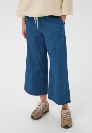 Personne portant un pantalon large en jean bleu, un sweat-shirt beige et des baskets marron clair, debout sur un fond clair.