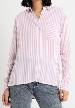 Femme portant une chemise à rayures verticales rose clair et blanche boutonnée, avec une poche poitrine rayée contrastante et un jean bleu clair.