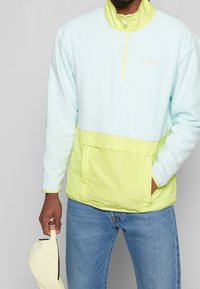 Pullover a mezza zip di colore azzurro chiaro e verde neon, con una texture morbida, una grande tasca frontale e un colletto alto. Tessuto pesante.