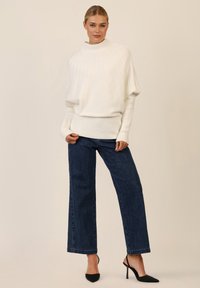 Übergroßer weißer gerippter Pullover mit hohem Kragen, kombiniert mit dunkelblauen weit geschnittenen Jeans mit dezenten Verzierungen und schwarzen spitzen Absätzen.
