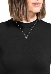 Silverfärgad trollslända hängsmycke på en tunn kedja, bärs över en svart höghalshalsad topp, som visar upp intrikata detaljer och design.