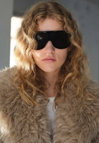 Occhiali da sole oversize neri, colletto in pelliccia marrone chiaro a trama, capelli biondi ondulati e colore delle labbra neutro. Top bianco semplice sotto.