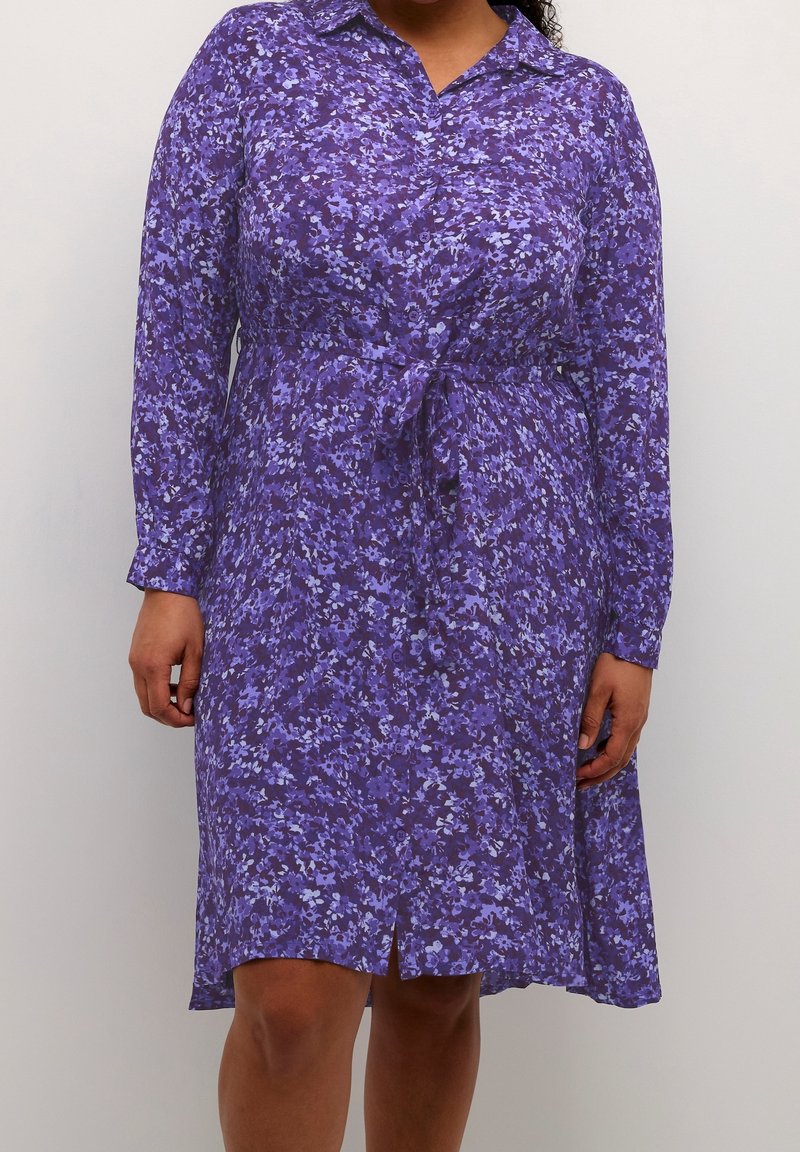 Femme portant une robe longue à manches longues à fleurs violettes avec des boutons et une ceinture nouée à la taille, debout devant un fond clair uni.