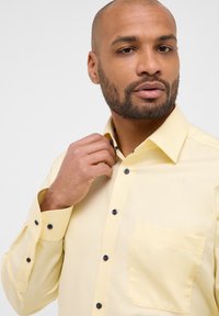 Camicia gialla con bottoni, caratterizzata da un sottile motivo testurizzato, bottoni scuri, una tasca sul petto e una fodera a scacchi all'interno del colletto.