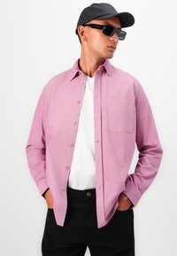 Chemise rose clair à boutons avec un tissu texturé, poche poitrine unique et manches longues, superposée sur un t-shirt blanc et un pantalon noir.